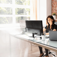 Person working at a modern office desk with dual monitors and a laptop, seated in front of a large bright window with sunlight streaming in, exposed brick wall and small plants in the background.