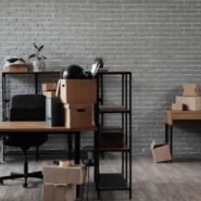 Office space in preparation for relocation, featuring stacked cardboard boxes, metal shelving with plants, and empty desks against a light brick wall.