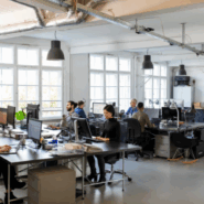 Modern open-plan office with a team of diverse professionals working at desks with multiple monitors, under industrial lighting and large windows.